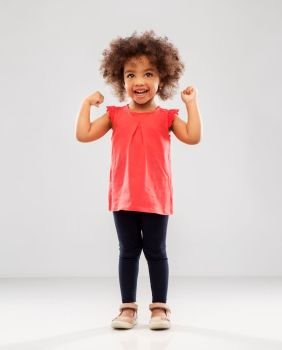 childhood and people concept - happy little african american girl showing her power over grey background. happy little african american girl showing power