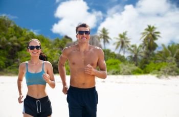 fitness, sport and lifestyle concept - happy couple in sports clothes and sunglasses running over tropical beach on island of seychelles background. couple in sports clothes running along on beach