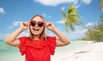 childhood, valentine’s day and summer concept - happy preteen girl with heart shaped sunglasses over tropical beach background in french polynesia. happy girl with heart shaped sunglasses on beach