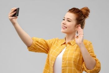 technology and people concept - smiling red haired teenage girl in checkered shirt taking selfie by smartphone and waving hand over grey background. redhead teenage girl taking selfie by smartphone