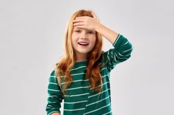 childhood, stress and people concept - happy smiling red haired girl in green striped shirt holding to her head over grey background. smiling red haired girl holding to her head