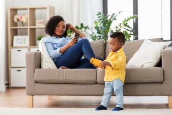 family, motherhood and people concept - african american baby playing with toy blocks kit and mother his using smartphone at home. baby playing toy car and mother with smartphone