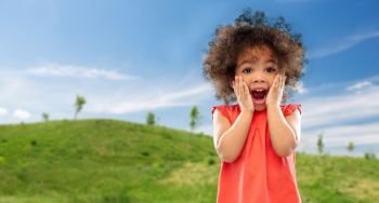 childhood, summer and emotions concept - surprised or scared little african american girl screaming over green meadow and blue sky background. surprised or scared little african american girl