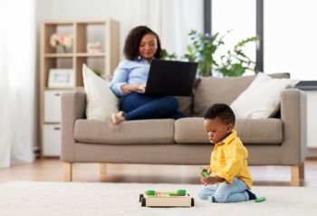 family, motherhood and people concept - african american baby playing with toy blocks kit and his mother using laptop computer at home. baby playing toy blocks and mother using laptop