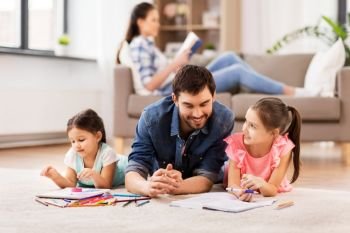family, fatherhood and leisure concept - father spending time with his little daughters drawing in sketchbooks by crayons and lying on floor at home. father with little daughters drawing at home