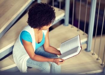 education, high school, university, learning and people concept - smiling african american student girl reading book sitting on stairs at library. african student girl reading book at library