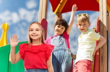 childhood, greeting and people concept - beautiful smiling girl in red shirt and skirt waving hand over children on outdoor kids playground background. smiling girl waving hand on kids playground