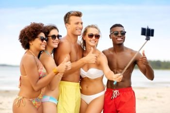 friendship, summer holidays and people concept - group of happy friends taking picture by smartphone on selfie stick on beach and showing thumbs up. friends taking selfie on beach and show thumbs up