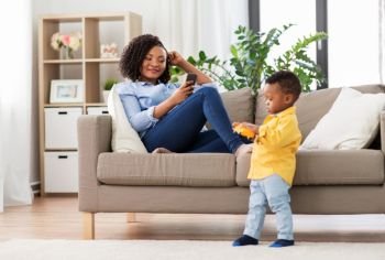 family, motherhood and people concept - african american mother using smartphone and baby playing toy car at home. mother using smartphone and baby playing toy car
