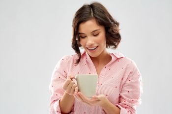 people and bedtime concept - happy young woman in pajama drinking coffee from mug over grey background. young woman in pajama drinking coffee from mug
