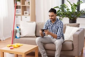 household and technology concept - indian man playing game on smartphone after cleaning home. man playing game on smartphone after cleaning home