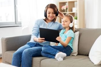 people, family and technology concept - happy mother and daughter with tablet pc computer and headphones listening to music at home. mother and daughter listen to music on tablet pc