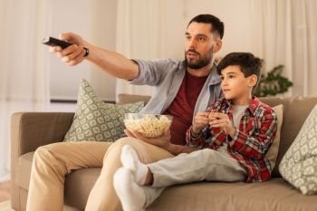 family, childhood, fatherhood, technology and people concept - father and little son with popcorn and remote control watching tv at home in evening. father and son with popcorn watching tv at home