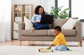 family, motherhood and people concept - happy african american mother using laptop computer and little baby son playing with toy blocks kit at home. mother using laptop and baby playing toy blocks