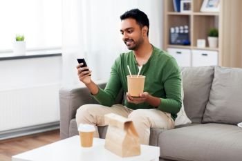 communication, leisure and people concept - smiling indian man using smartphone and eating takeaway food at home. smiling indian man eating takeaway food at home