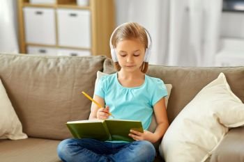 people, childhood and bedtime concept - little girl in headphones with diary and pencil sitting on sofa at home. girl in headphones with diary on sofa at home