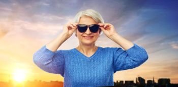 valentine’s day, summer and old people concept - portrait of smiling senior woman in red black sunglasses over sunset in tallinn city background. senior woman in sunglasses over sunset in city