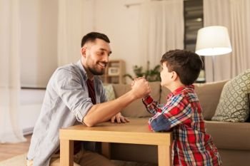 family, fatherhood and people concept - happy smiling father and little son arm wrestling at home in evening. happy father and little son arm wrestling at home