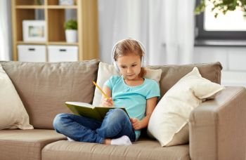 people, childhood and bedtime concept - little girl in headphones with diary and pencil sitting on sofa at home. girl in headphones with diary on sofa at home
