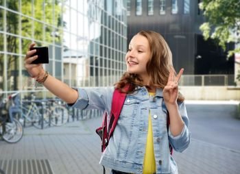 education and people concept - happy smiling teenage student girl with bag taking selfie by smartphone and showing peace over school yard background. teenage student girl taking selfie by smartphone