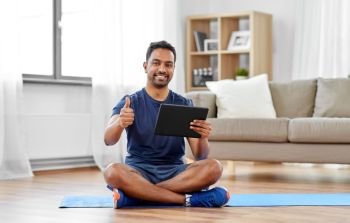 sport, technology and healthy lifestyle concept - smiling indian man with tablet computer sitting on exercise mat and showing thumbs up at home. indian man with tablet computer on exercise mat