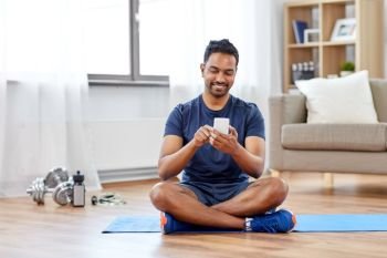 sport, technology and healthy lifestyle concept - smiling indian man with smartphone sitting on exercise mat at home. indian man with smartphone on exercise mat at home