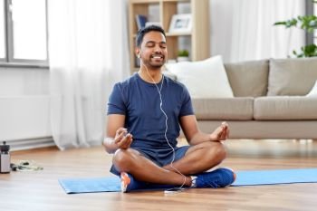 fitness, meditation and healthy lifestyle concept - indian man in earphones listening to music on smartphone and meditating in lotus pose at home. indian man meditating in lotus pose at home
