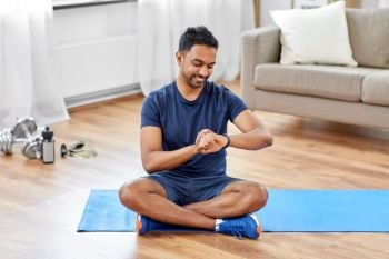 sport, technology and healthy lifestyle concept - smiling indian man with fitness tracker sitting on exercise mat at home. smiling indian man with fitness tracker at home