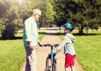 family, generation, safety and people concept - happy grandfather and boy with bicycle and bike helmet talking at summer park. grandfather and boy with bicycle at summer park