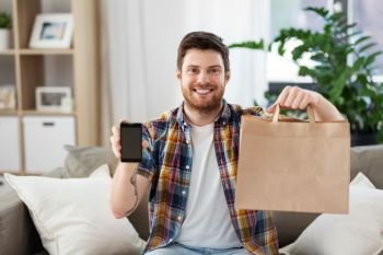 technology, consumption and people concept - smiling man showing smartphone and food delivery bag at home. man using smartphone for food delivery