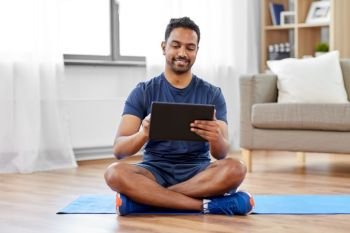 sport, technology and healthy lifestyle concept - smiling indian man with tablet computer sitting on exercise mat at home. indian man with tablet pc and exercise mat at home