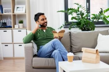 communication, leisure and people concept - smiling indian man calling on smartphone and eating takeaway food at home. smiling indian man eating takeaway food at home