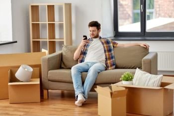 moving, people and real estate concept - happy man with smartphone and boxes sitting on sofa at new home. man with smartphone and boxes on sofa at new home