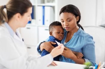 medicine, healthcare and pediatry concept - african american mother with sick baby son and caucasian doctor with thermometer at clinic. mother with sick baby son and doctor at clinic