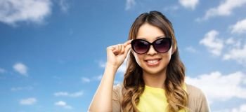 summer fashion, style and eyewear concept - happy smiling young asian woman in sunglasses over blue sky and clouds background. happy smiling young asian woman in sunglasses