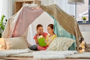 childhood, hygge and friendship concept - happy girls reading book with torch light in kids tent or teepee at home. happy girls reading book in kids tent at home