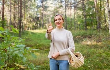 season and leisure people concept - young woman with mushrooms in wicker basket in forest showing thumbs up. woman with basket of mushrooms in forest