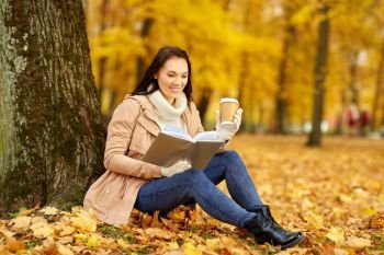season, technology and people concept - young woman reading book and drinking takeaway coffee from paper cup in autumn park. woman reading book with coffee in autumn park