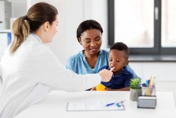 medicine, healthcare and pediatry concept - caucasian doctor with stick examining african american baby boy patient’s mouth at clinic. doctor examining african baby’s mouth at clinic