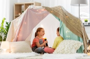 childhood, hygge and friendship concept - happy girls playing with torch light in kids tent at home. girls playing with torch in kids tent at home