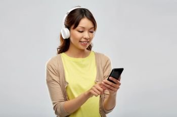 people, technology and audio equipment concept - happy asian young woman in headphones listening to music on smartphone over grey background. asian woman in headphones listening to music