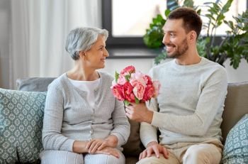 family, mother’s day and birthday concept - smiling adult son giving flowers to his senior mother at home. adult son giving flowers to senior mother at home