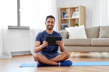 sport, technology and healthy lifestyle concept - smiling indian man with tablet computer sitting on exercise mat at home. indian man with tablet pc and exercise mat at home