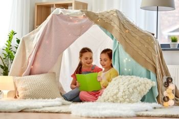childhood, hygge and friendship concept - happy girls reading book in kids tent or teepee at home. happy girls reading book in kids tent at home