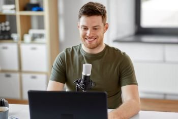 technology, mass media and podcast concept - happy young male audio blogger with laptop computer and microphone broadcasting at home office. man with laptop and microphone at home office