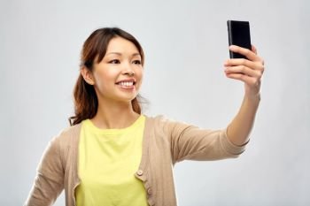 technology and people concept - happy asian woman taking selfie by smartphone over grey background. asian woman taking selfie by smartphone