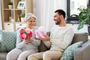 family, mother’s day and birthday concept - smiling adult son giving present and flowers to his senior mother at home. son giving present and flowers to senior mother