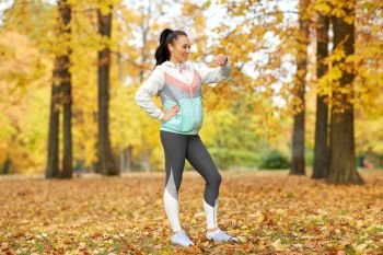 sport, technology and healthy lifestyle concept - young woman with earphones and fitness tracker in autumn park. woman looking at fitness tracker in autumn park