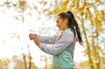sport, technology and healthy lifestyle concept - young woman with earphones and fitness tracker in autumn park. woman looking at fitness tracker in autumn park