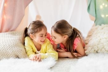 childhood, hygge and friendship concept - happy girls lying in kids tent and talking at home. happy girls lying in kids tent and talking at home